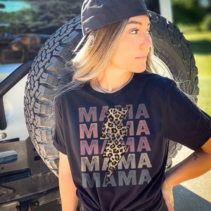 Op de afbeelding: Een zwart T-shirt met een afbeelding van een bliksemschicht met luipaardprint en het woord "MAMA" dat in een vervaagd, gelaagd ontwerp wordt herhaald. Het shirt wordt gedragen door een persoon met blond haar en een zwarte baseballpet.