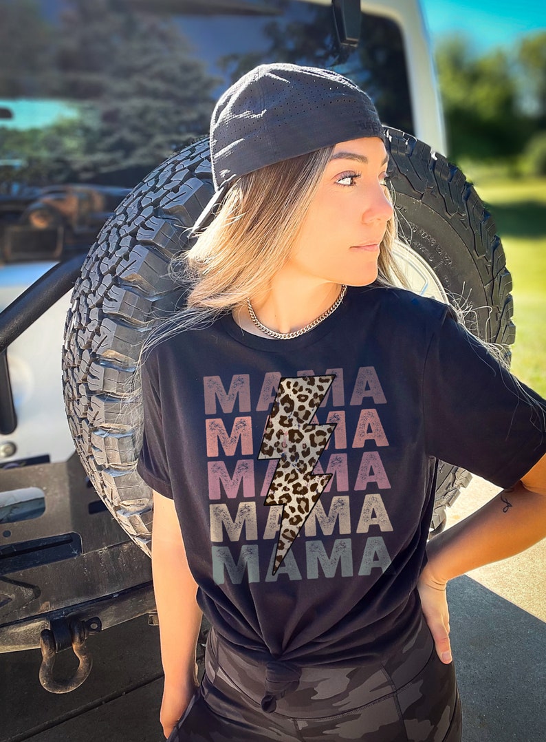 Op de afbeelding: Een zwart T-shirt met een afbeelding van een bliksemschicht met luipaardprint en het woord "MAMA" dat in een vervaagd, gelaagd ontwerp wordt herhaald. Het shirt wordt gedragen door een persoon met blond haar en een zwarte baseballpet.
