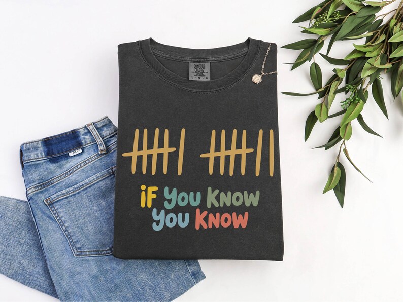 May include: Dark gray t-shirt with the text "if you know you know" in colorful letters. The shirt also features a tally mark design in gold. A pair of blue jeans is next to the shirt.