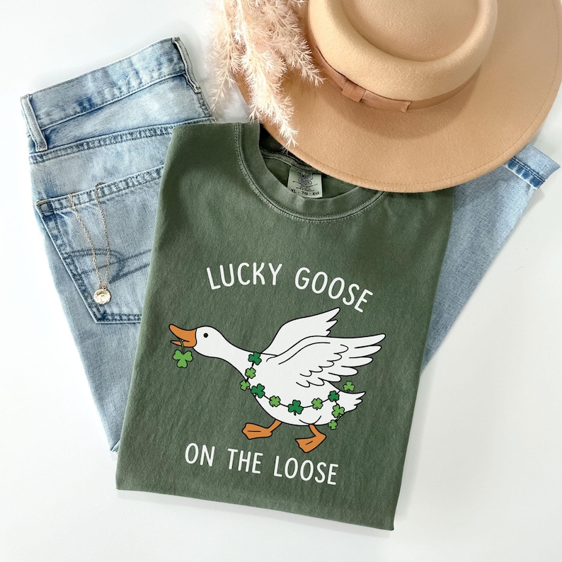 May include: An olive green t-shirt with the text "LUCKY GOOSE ON THE LOOSE" and a cartoon goose wearing a shamrock garland. The shirt is paired with light wash jeans and a tan hat.
