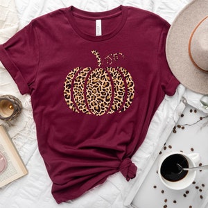 May include: A maroon t-shirt with a leopard print pumpkin design. The shirt is laid out on a white surface with a coffee cup, a book, and a hat.