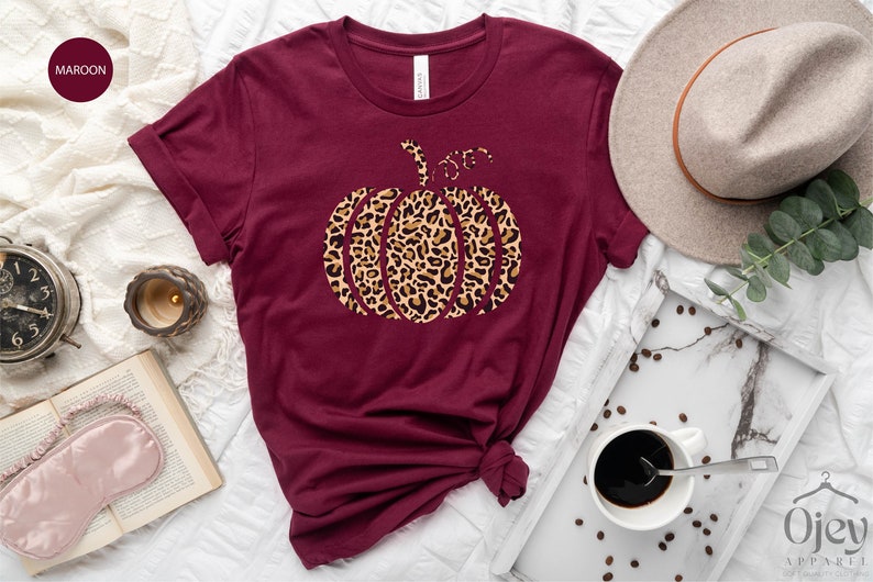 May include: A maroon t-shirt with a leopard print pumpkin design. The shirt is laid out on a white surface with a coffee cup, a book, and a hat.