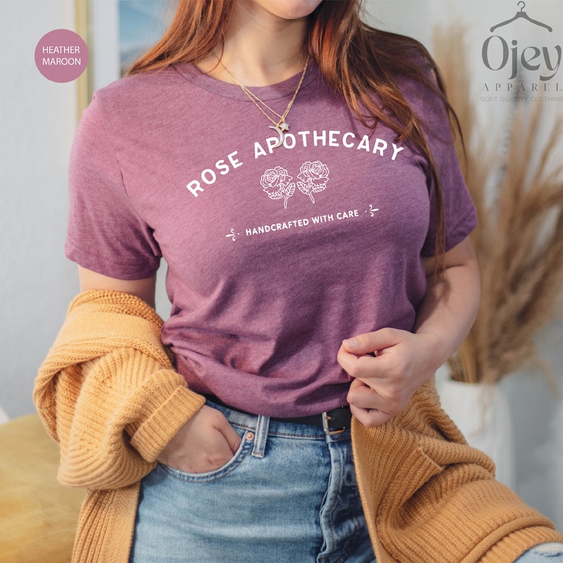 May include: Heather maroon t-shirt with the text "ROSE APOTHECARY" in white, along with two rose illustrations and the phrase "HANDCRAFTED WITH CARE." The shirt is paired with blue jeans and a mustard-colored cardigan.