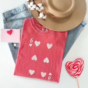 May include: A coral-colored t-shirt with white heart graphics and the number 6. The shirt is paired with light blue jeans, a tan hat, a pink card with a red heart, and a heart-shaped lollipop.