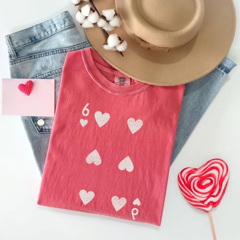 May include: A coral-colored t-shirt with white heart graphics and the number 6. The shirt is paired with light blue jeans, a tan hat, a pink card with a red heart, and a heart-shaped lollipop.