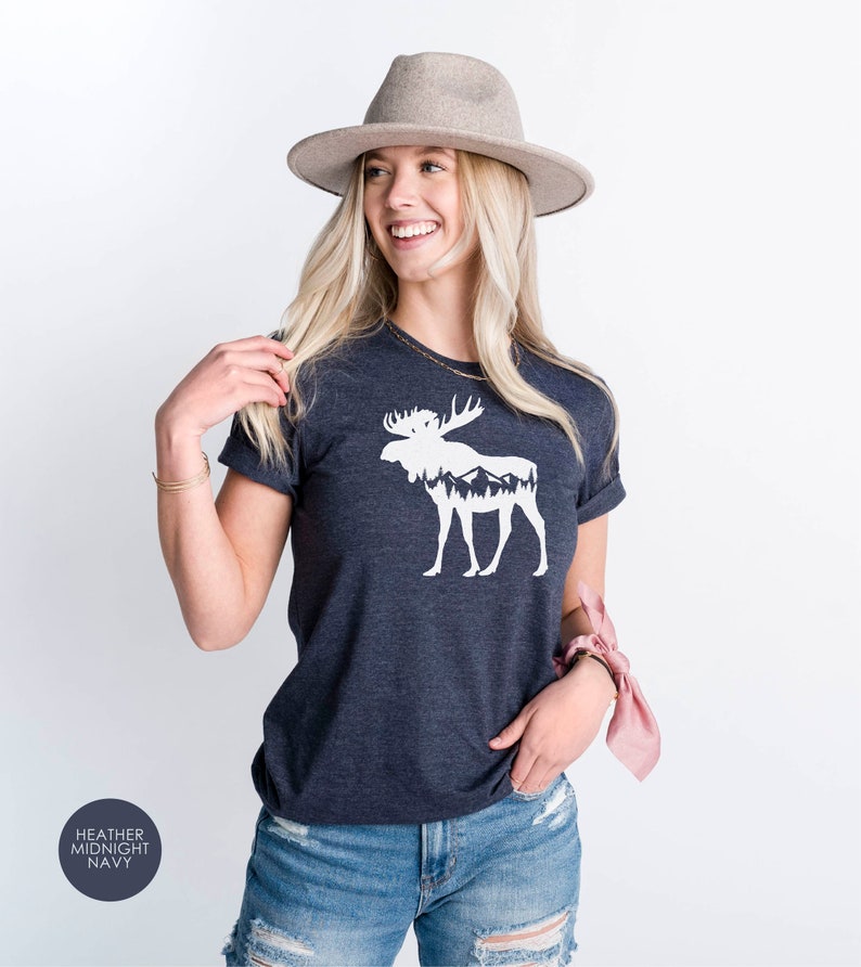 May include: Heather midnight navy t-shirt featuring a white moose graphic with mountain and forest details. The shirt has rolled sleeves and a crew neck. The text "Heather Midnight Navy" is in a circle in the lower left corner.