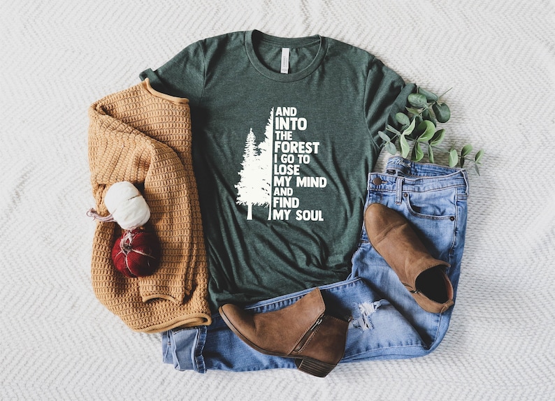 May include: A green t-shirt with a white tree and the text "And into the forest I go to lose my mind and find my soul". The shirt is laid out on a white fuzzy surface with a brown sweater, blue jeans, and brown boots.