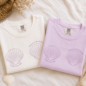 May include: Two crew neck t-shirts, one white and one light purple, each featuring two purple line-art seashell illustrations. The text "Jasmine's Bachelorette" and "Punta Cana 2026" is printed in purple near the shells on both garments.