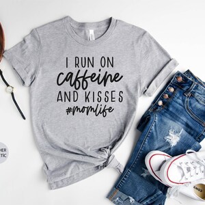 K&ouml;nnte beinhalten: Graues T-Shirt mit dem Text "I run on caffeine and kisses #momlife" in Schwarz gedruckt.