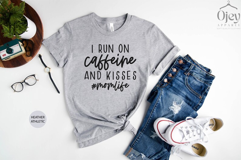 K&ouml;nnte beinhalten: Graues T-Shirt mit dem Text "I run on caffeine and kisses #momlife" in Schwarz gedruckt.