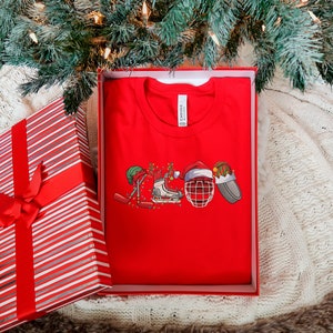 May include: Red t-shirt with a Christmas-themed hockey design. The design features a hockey stick, skates, a hockey helmet, and a Santa hat.