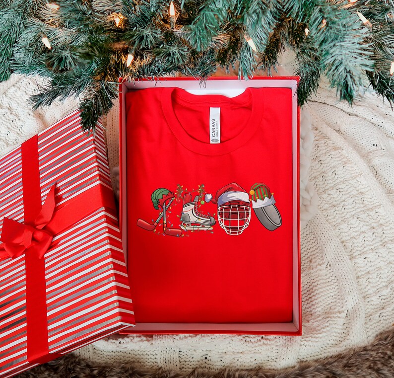May include: Red t-shirt with a Christmas-themed hockey design. The design features a hockey stick, skates, a hockey helmet, and a Santa hat.