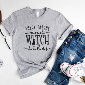 May include: Gray heather athletic t-shirt with the text "Thick Thighs and Watch Vibes" in black lettering. The shirt is styled with blue jeans and white sneakers.