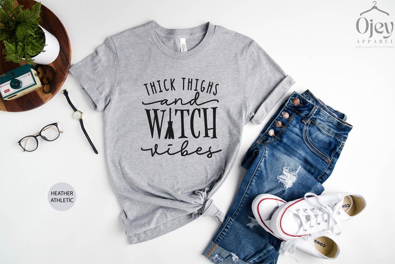 May include: Gray heather athletic t-shirt with the text "Thick Thighs and Watch Vibes" in black lettering. The shirt is styled with blue jeans and white sneakers.