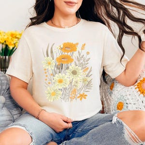 K&ouml;nnte beinhalten: Eine Frau tr&auml;gt ein cremefarbenes T-Shirt mit einem Blumendruck aus gelben und orangefarbenen Blumen. Das Shirt hat eine lockere Passform und kurze &Auml;rmel.
