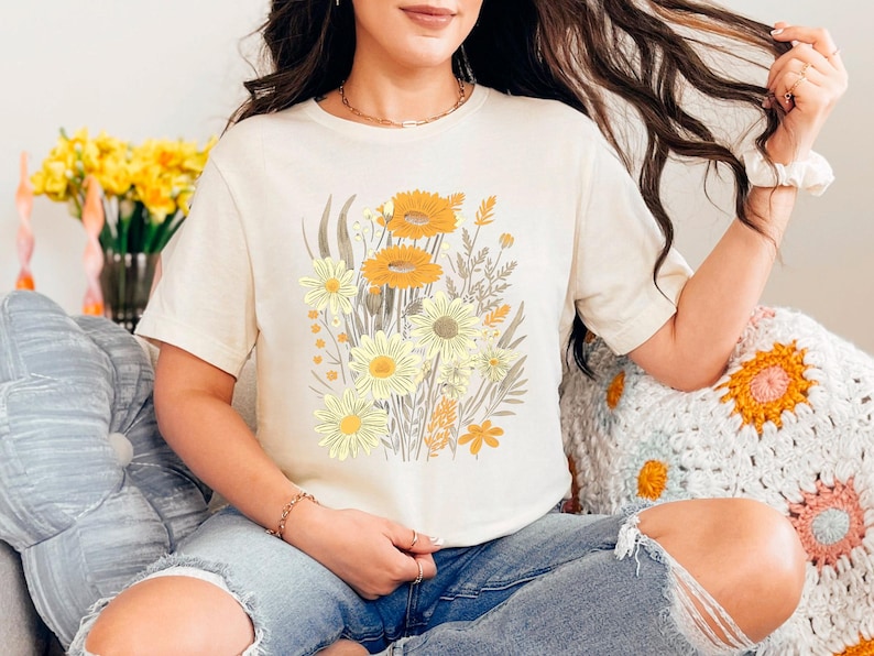 K&ouml;nnte beinhalten: Eine Frau tr&auml;gt ein cremefarbenes T-Shirt mit einem Blumendruck aus gelben und orangefarbenen Blumen. Das Shirt hat eine lockere Passform und kurze &Auml;rmel.