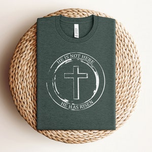 K&ouml;nnte beinhalten: Ein dunkelgr&uuml;nes T-Shirt mit einem wei&szlig;en Kreis-Motiv, das ein Kreuz und den Text "Er ist nicht hier, Er ist auferstanden" zeigt.