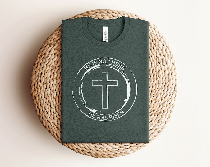 K&ouml;nnte beinhalten: Ein dunkelgr&uuml;nes T-Shirt mit einem wei&szlig;en Kreis-Motiv, das ein Kreuz und den Text "Er ist nicht hier, Er ist auferstanden" zeigt.