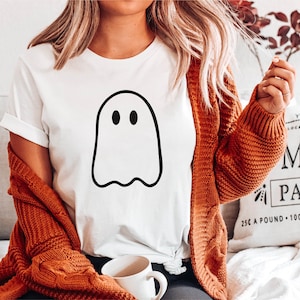 May include: White t-shirt with a black outline of a ghost with two eyes. The shirt is being worn by a person with long blonde hair and a brown cardigan. The person is sitting on a couch with a white pillow that says "Your Own Locally Grown Pumpkin Patch 25&cent; a pound 100% Organic Open"
