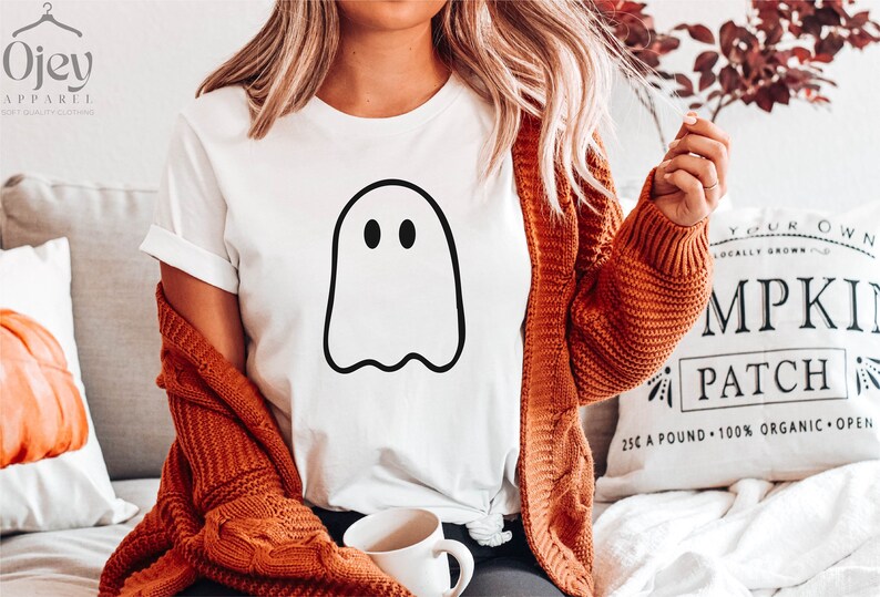May include: White t-shirt with a black outline of a ghost with two eyes. The shirt is being worn by a person with long blonde hair and a brown cardigan. The person is sitting on a couch with a white pillow that says "Your Own Locally Grown Pumpkin Patch 25&cent; a pound 100% Organic Open"