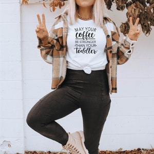 May include: A woman wearing a white t-shirt with the text "May your coffee be stronger than your toddler" printed on it. She is wearing a brown plaid shirt over the t-shirt, black leggings, and beige boots. She is standing on a tiled floor with a star pattern.