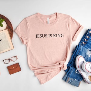 Pode incluir: Uma camiseta cor p&ecirc;ssego com o texto "JESUS IS KING" estampado na frente. A camiseta est&aacute; colocada sobre uma superf&iacute;cie branca com um par de cal&ccedil;as jeans, t&ecirc;nis brancos, um livro, uma caneta, &oacute;culos e uma carteira marrom.