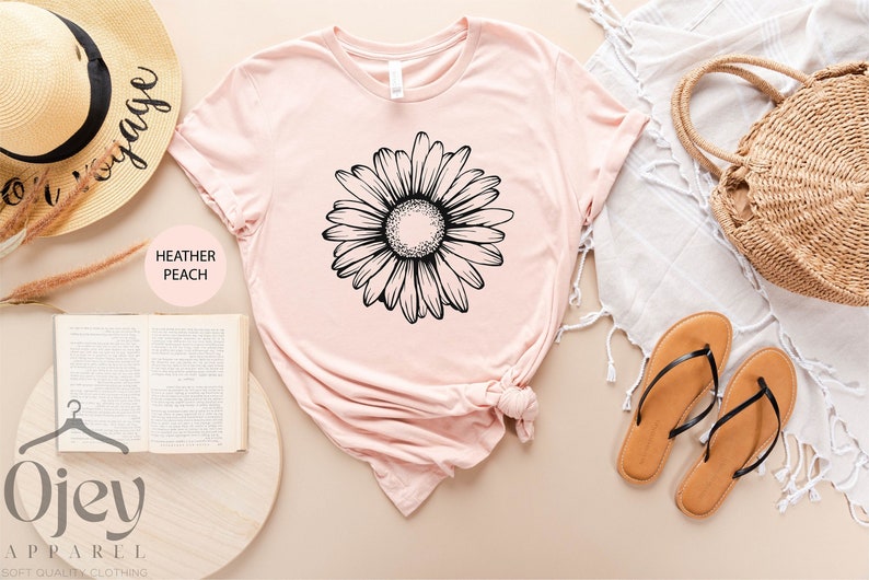 Puede incluir: Una camiseta de color melocot&oacute;n jaspeado con un gr&aacute;fico de margarita en blanco y negro. La camiseta est&aacute; colocada sobre una superficie marr&oacute;n claro con un sombrero de paja, un libro, un bolso tejido y unas sandalias.