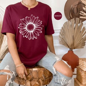 Puede incluir: Una camiseta de color granate con un gr&aacute;fico de margarita blanca. La camiseta la lleva puesta una persona que lleva pantalones vaqueros azules.