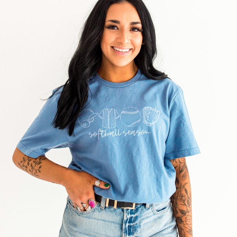 May include: A light blue t-shirt with a white graphic of a baseball helmet, jersey, ball, and glove. The text "softball season" is printed below the graphic.