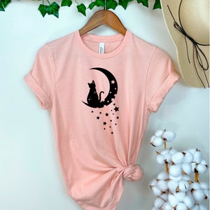 May include: A pink t-shirt with a black silhouette of a cat sitting on a crescent moon with stars. The shirt is knotted at the bottom.