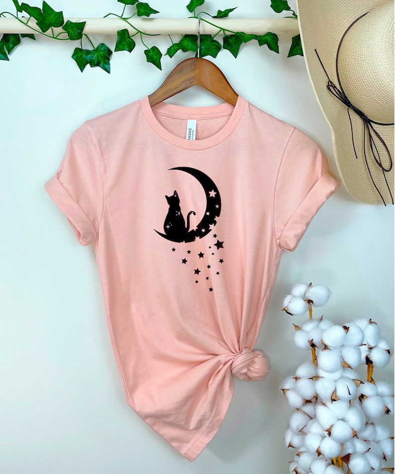 May include: A pink t-shirt with a black silhouette of a cat sitting on a crescent moon with stars. The shirt is knotted at the bottom.