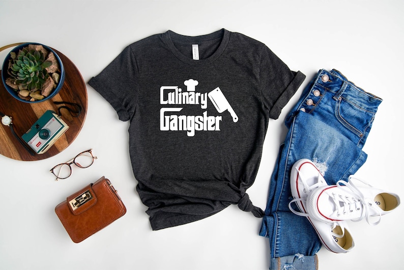 May include: A dark gray t-shirt with the text "Culinary Gangster" in white lettering. The text is accompanied by a chef's hat and a white silhouette of a butcher knife.