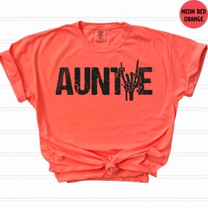May include: Neon red-orange t-shirt with the word "AUNTIE" in distressed black lettering and a skeleton hand making a rock and roll gesture. The shirt is knotted at the waist and has rolled-up sleeves.
