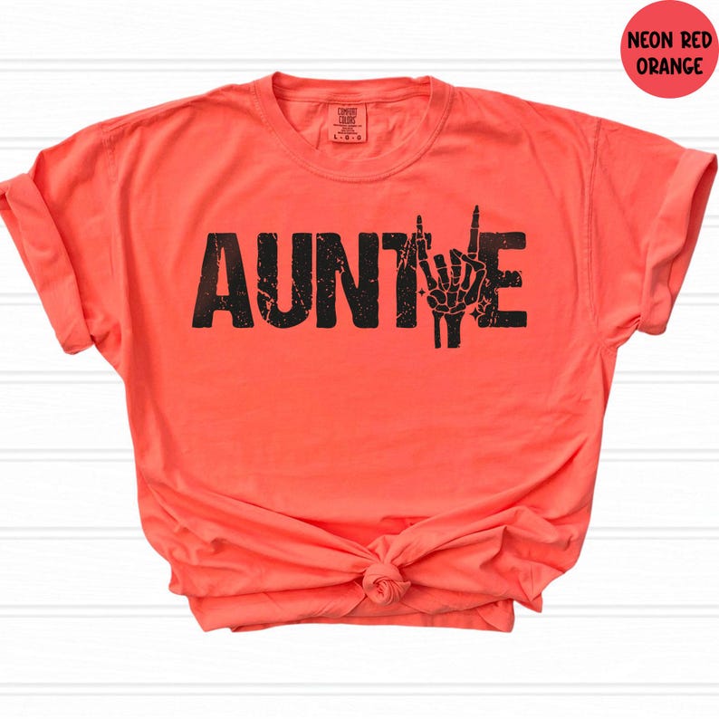 May include: Neon red-orange t-shirt with the word "AUNTIE" in distressed black lettering and a skeleton hand making a rock and roll gesture. The shirt is knotted at the waist and has rolled-up sleeves.