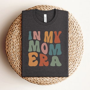 May include: A dark gray t-shirt with the text "IN MY MOM ERA" in colorful, retro-style lettering.