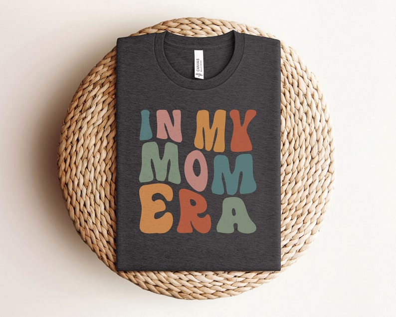 May include: A dark gray t-shirt with the text "IN MY MOM ERA" in colorful, retro-style lettering.