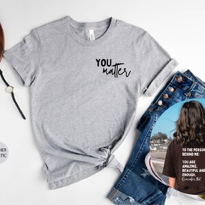 May include: A heather grey t-shirt with the text "YOU matter" printed on the front. The shirt is shown on a white surface with a pair of jeans, a watch, and a camera.