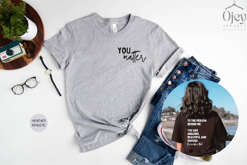 May include: A heather grey t-shirt with the text "YOU matter" printed on the front. The shirt is shown on a white surface with a pair of jeans, a watch, and a camera.
