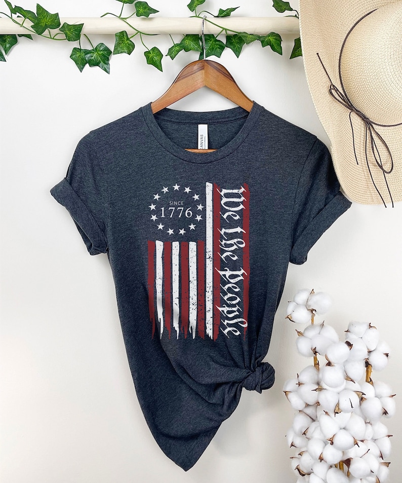 May include: A dark gray t-shirt with a distressed American flag design and the text "We the People" and "Since 1776" printed on it.