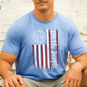 May include: A light blue t-shirt with a distressed American flag design. The flag has red and white stripes and a blue field with white stars. The text "We the People" is written in a cursive font on the right side of the flag. The text "Since 1776" is written in a white font on the blue field of the flag.
