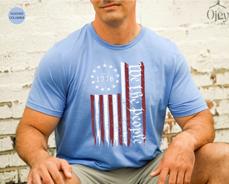 May include: A light blue t-shirt with a distressed American flag design. The flag has red and white stripes and a blue field with white stars. The text "We the People" is written in a cursive font on the right side of the flag. The text "Since 1776" is written in a white font on the blue field of the flag.