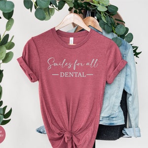K&ouml;nnte beinhalten: Ein T-Shirt in der Farbe Heide-Himbeere mit dem Text "Smiles for all - DENTAL" in wei&szlig;en Buchstaben.