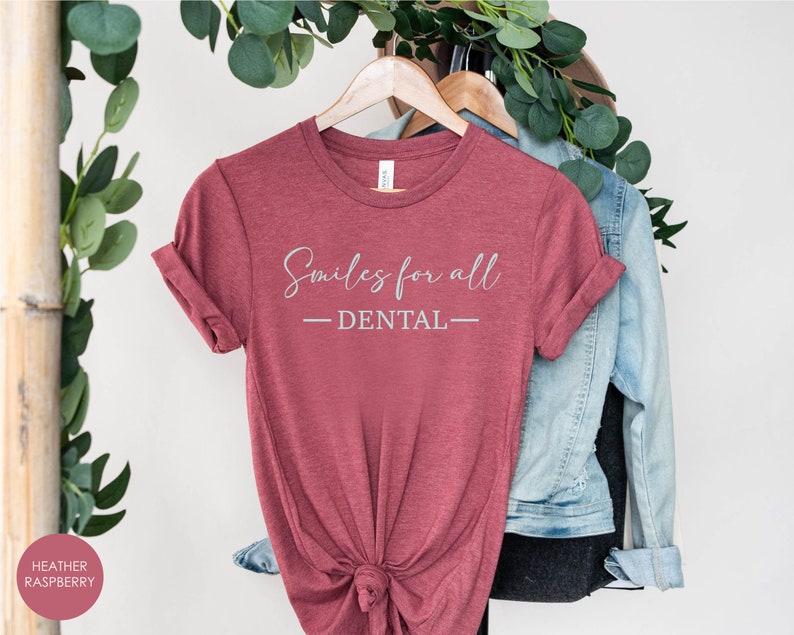 K&ouml;nnte beinhalten: Ein T-Shirt in der Farbe Heide-Himbeere mit dem Text "Smiles for all - DENTAL" in wei&szlig;en Buchstaben.