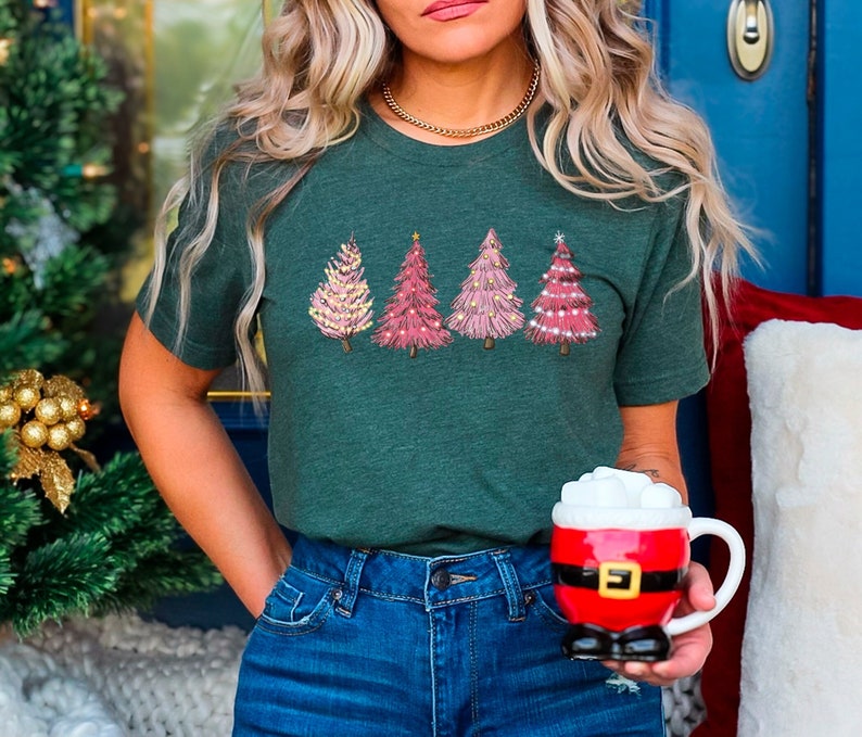 May include: A woman wearing a green t-shirt with a pink and white Christmas tree design. She is holding a red Santa mug with white marshmallows.