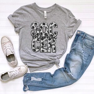 May include: Gray t-shirt with a black and white soccer ball design that says "Soccer Mom" on it. The shirt is paired with blue jeans and white sneakers.