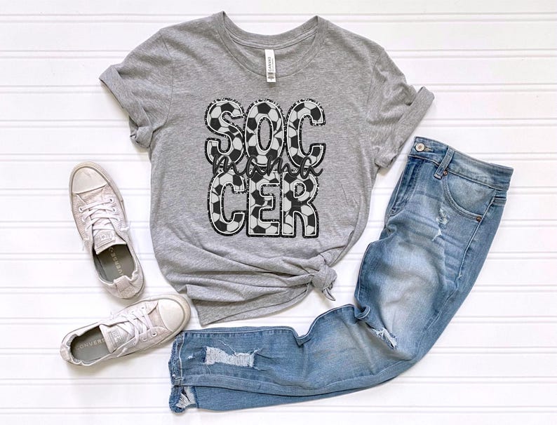 May include: Gray t-shirt with a black and white soccer ball design that says "Soccer Mom" on it. The shirt is paired with blue jeans and white sneakers.
