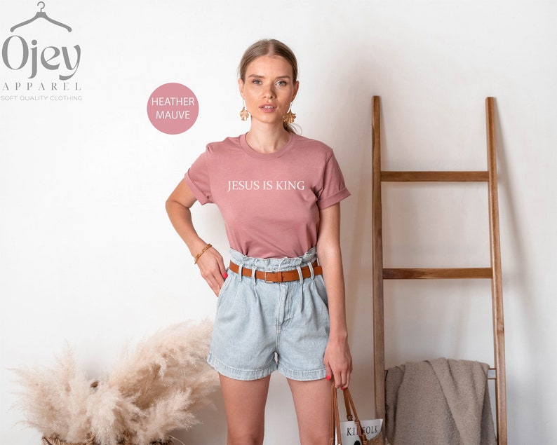 Pode incluir: Uma mulher vestindo uma camiseta malva com o texto "JESUS IS KING" em branco. Ela tamb&eacute;m est&aacute; vestindo shorts jeans azul claro com um cinto marrom e segurando uma bolsa de palha.