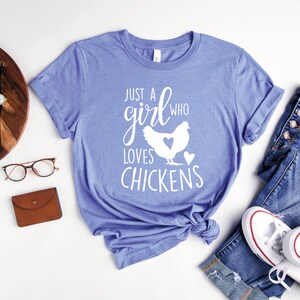 Puede incluir: Una camiseta azul con el texto "Just a girl who loves chickens" en letras blancas. La camiseta presenta una silueta blanca de una gallina con un coraz&oacute;n en el pecho.