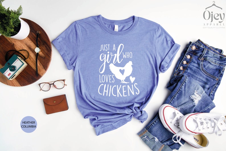 Puede incluir: Una camiseta azul con el texto "Just a girl who loves chickens" en letras blancas. La camiseta presenta una silueta blanca de una gallina con un coraz&oacute;n en el pecho.