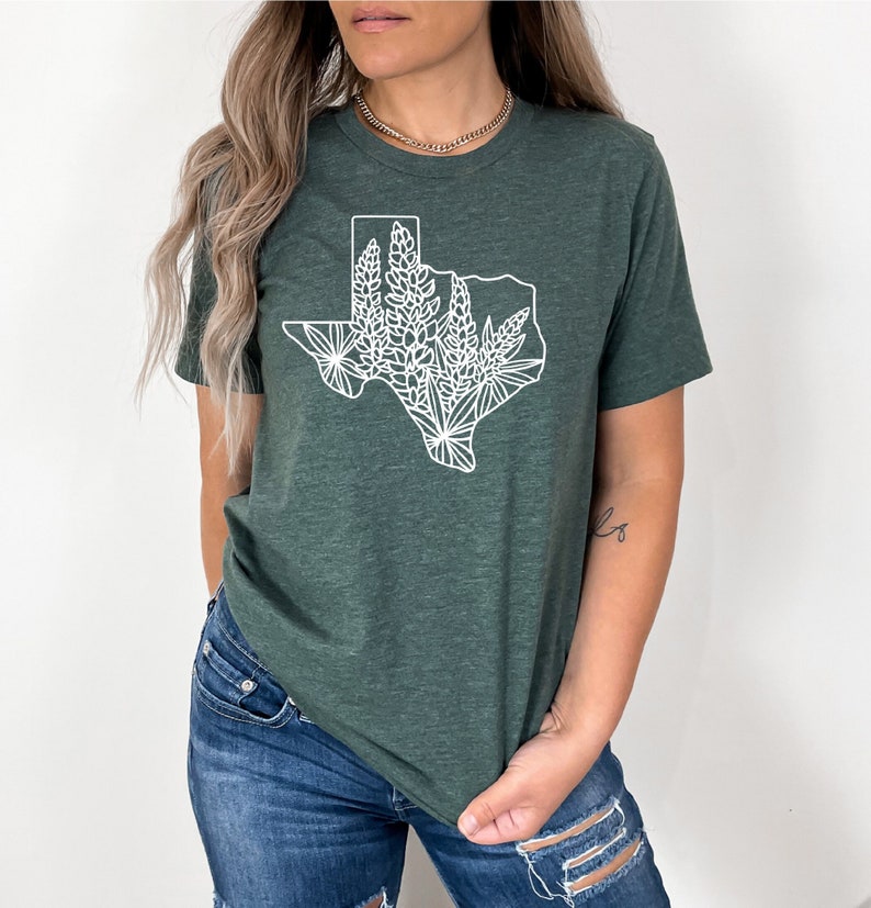 May include: Dark green t-shirt featuring a white outline of the state of Texas filled with floral designs. The shirt has short sleeves and a crew neck. The fabric appears to be a soft, heathered material.